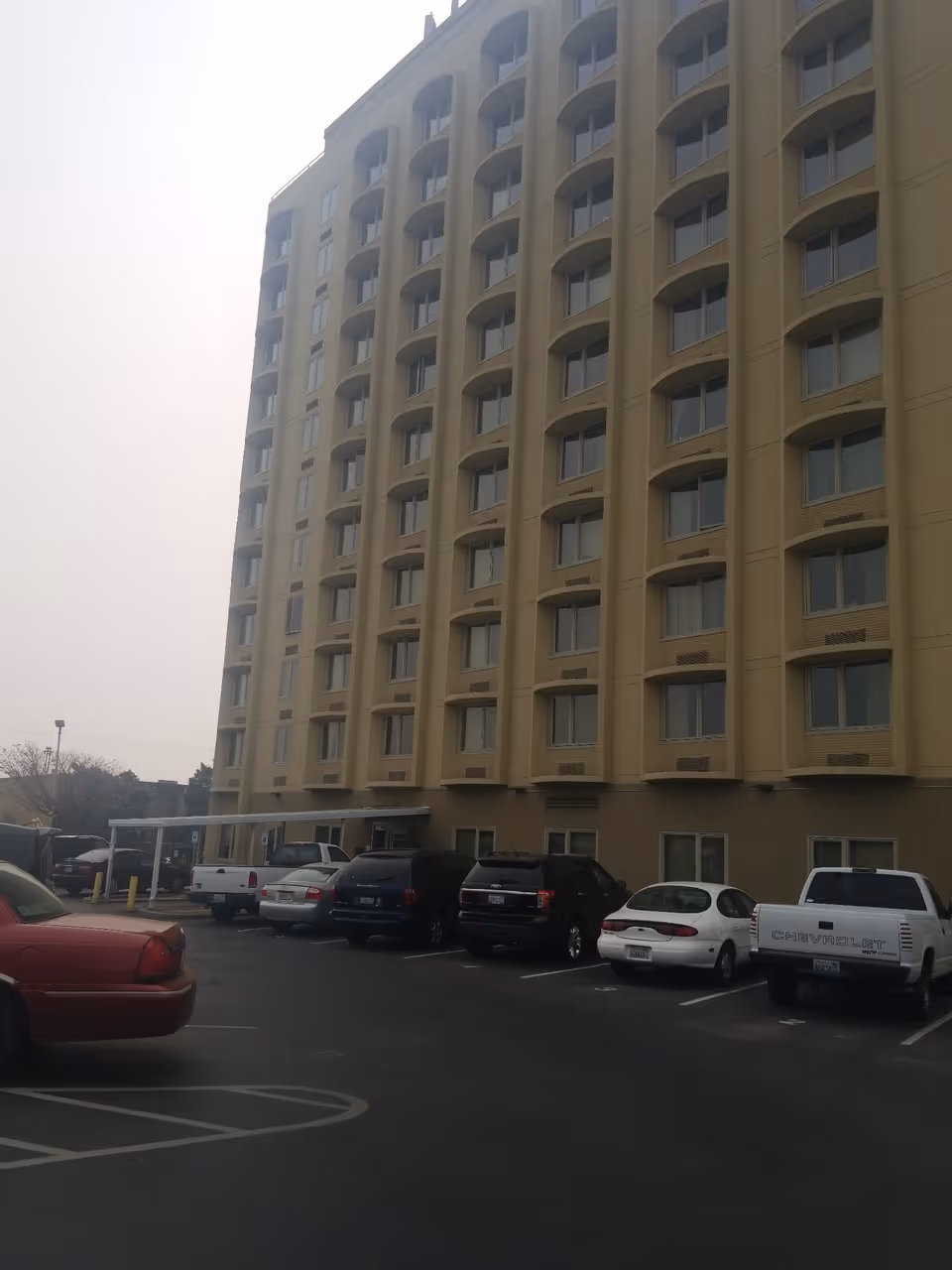 Exterior view of a multi-story beige building with many windows, likely a senior living facility, with a parking lot in front containing several parked cars and trucks under an overcast sky.
