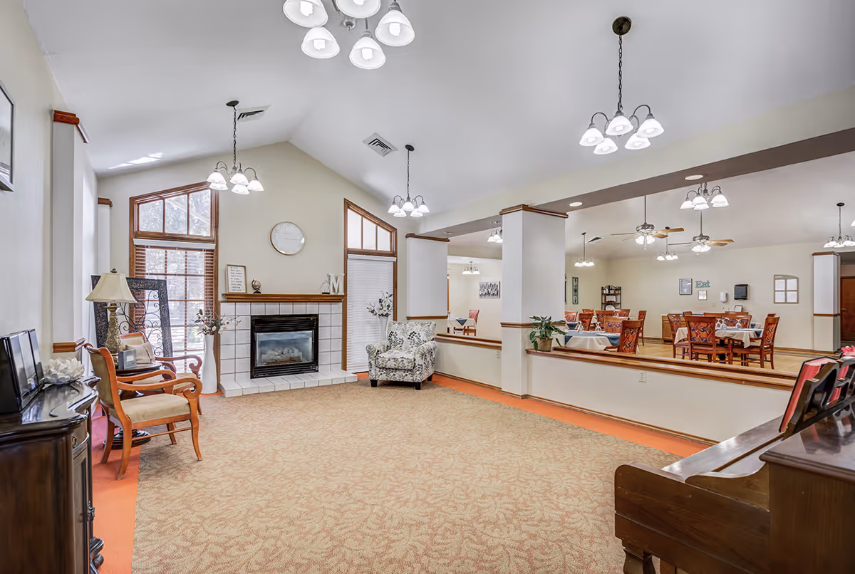A spacious senior living facility common area with beige carpet and white walls. The room features a fireplace with a clock above it, two large windows with wooden blinds, and several chairs including an armchair and wooden chairs. Multiple ceiling light fixtures illuminate the space. In the background, there is a dining area with tables and chairs set for meals.