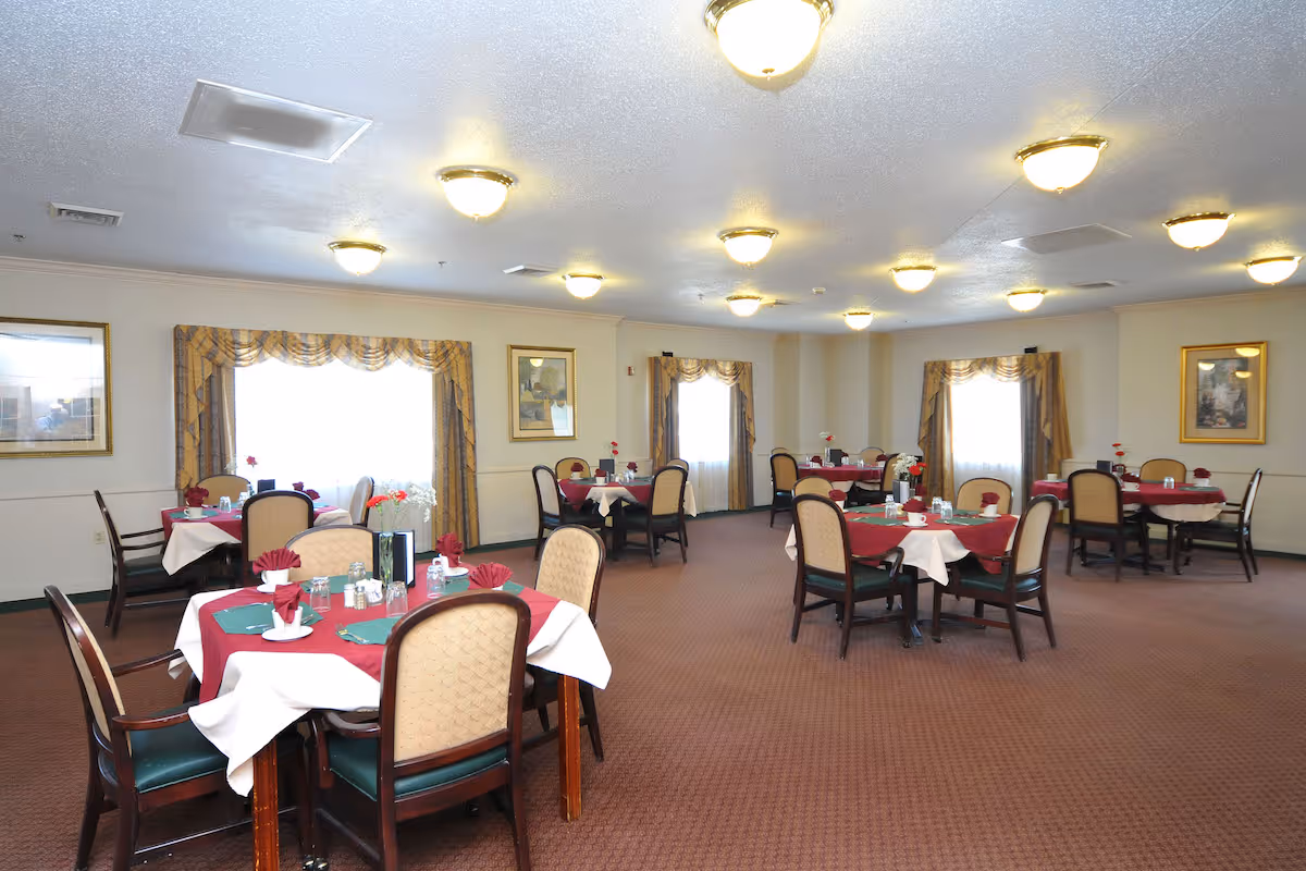 Large dining room with multiple round tables draped in red and white linens, place settings, and upholstered chairs under ceiling lights.