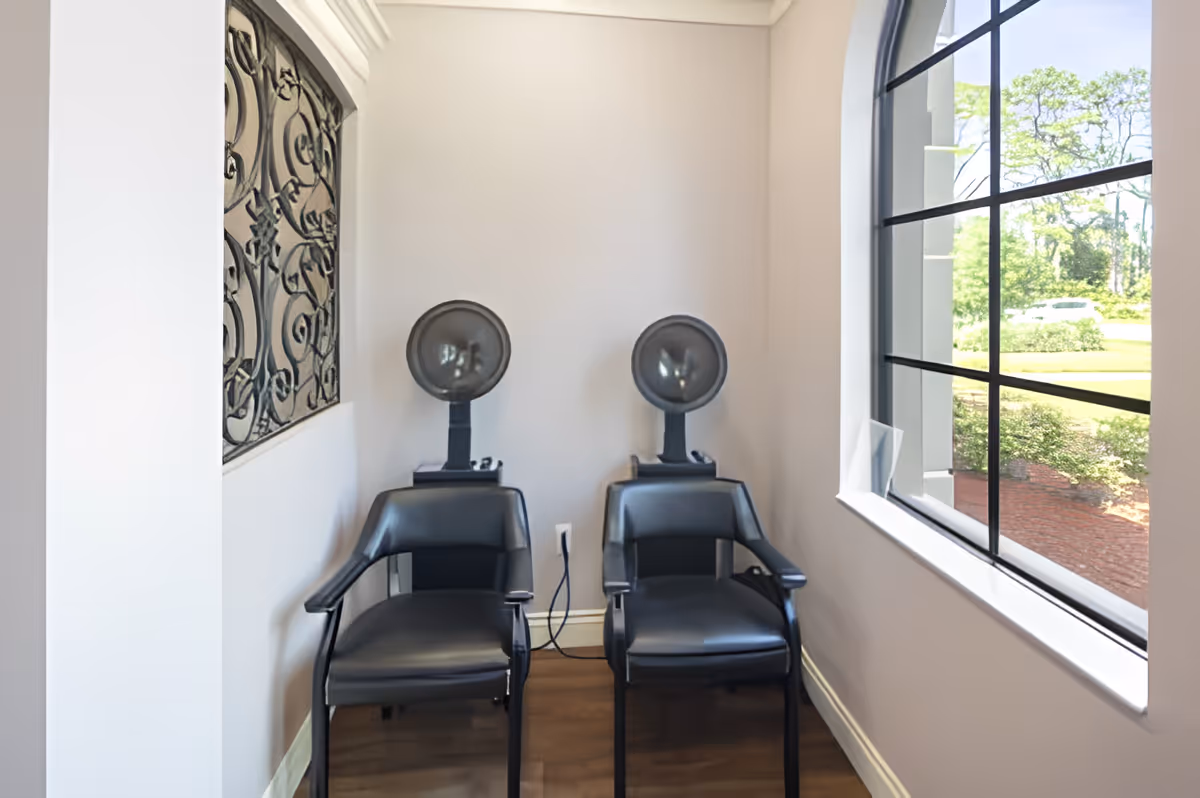 Two black salon hair dryer chairs positioned side by side in a small room with light-colored walls and a large window showing greenery outside. There is decorative wrought iron wall art on the left wall.