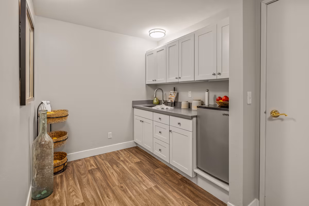 A small kitchen area with white cabinets, a gray countertop, a stainless steel mini refrigerator, and a sink. There are three woven baskets stacked on a metal frame on the left side, a large decorative glass bottle on the floor, and a bowl of red apples on the countertop. The floor has wood-style vinyl planks, and the walls are painted light gray.