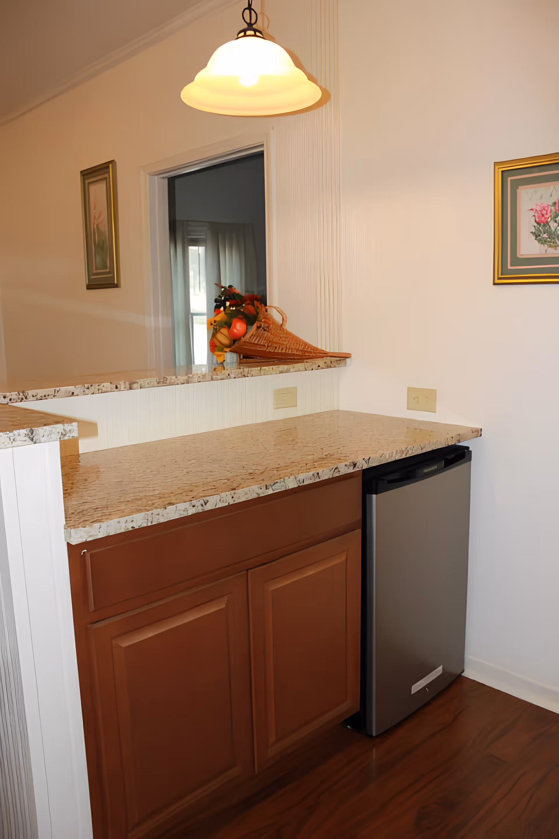 Kitchen pass-through with a granite countertop, wooden cabinets, a stainless mini-fridge, pendant light, and a decorative fruit basket.