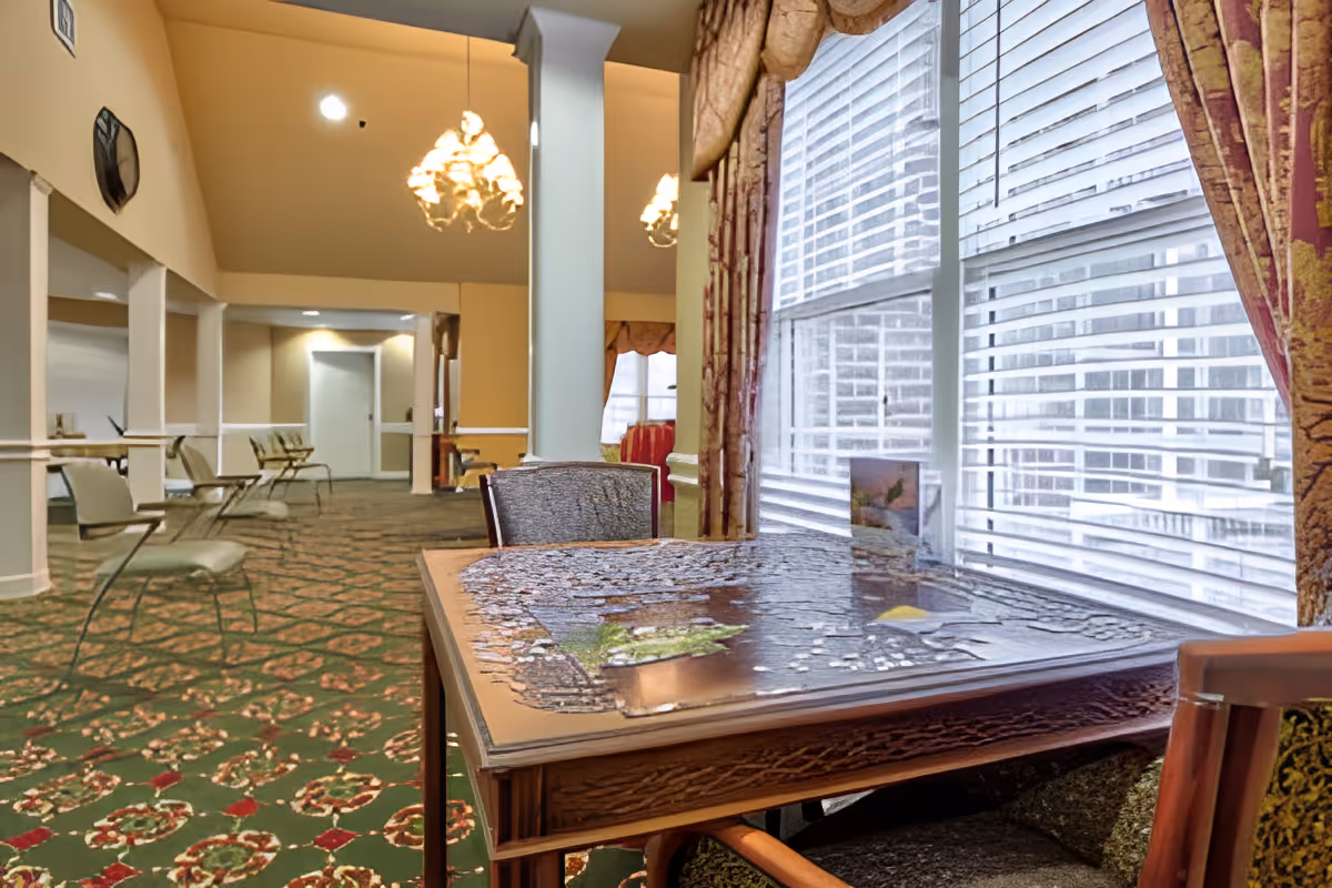 Sunlit common room with a table holding a partially completed jigsaw puzzle by a window, chairs, patterned carpet, and chandeliers.