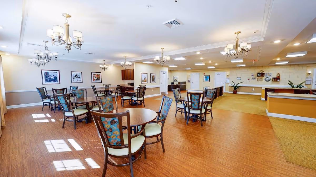 Spacious senior living dining room with round wooden tables, patterned upholstered chairs, chandeliers, and a reception desk in the background.