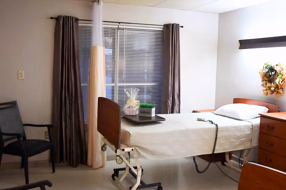 A nursing facility bedroom with a hospital bed covered in a white quilt, a pillow, and a remote control on the bed. There is a tray with a small plant and a gift basket on the bed. The room has a window with closed blinds and brown curtains, a black chair, a wooden dresser, and a decorative wreath on the wall.