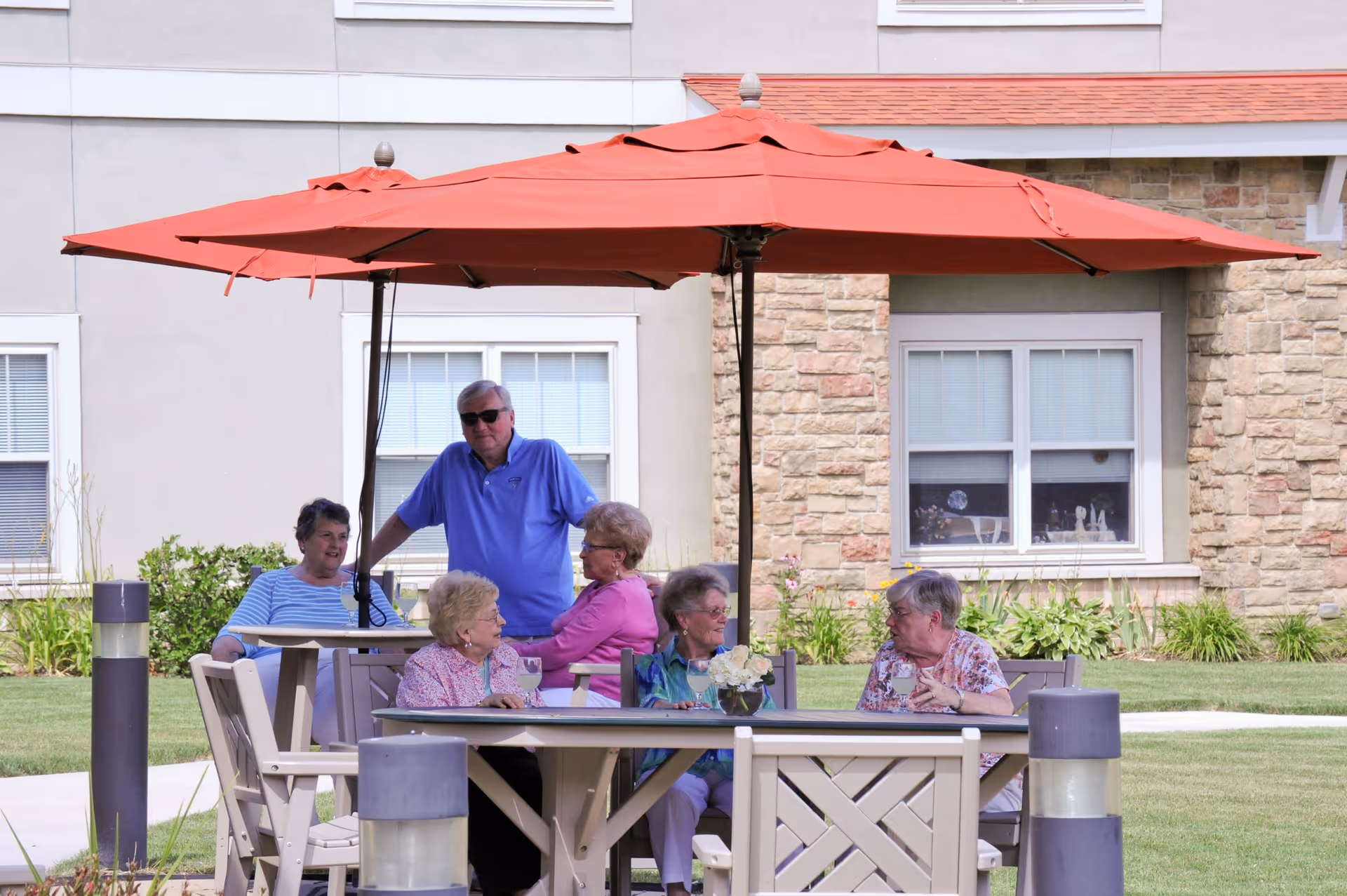 A group of elderly people sitting and standing around an outdoor table under a large red umbrella in a garden area of a senior living facility. The building with windows and stone facade is visible in the background.
