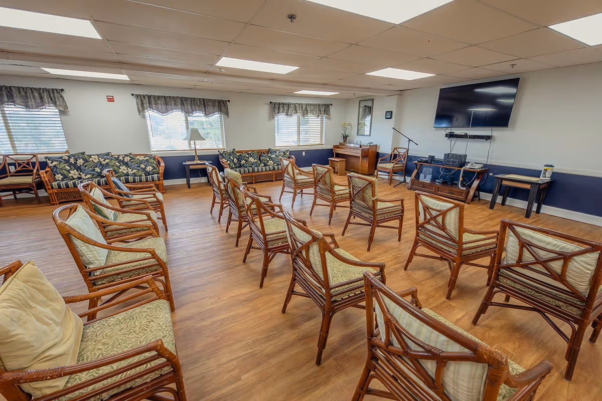 A spacious room with wooden flooring and multiple wooden chairs arranged in rows facing a wall-mounted flat screen TV. There are two floral-patterned sofas along the far wall beneath windows with valance curtains. A piano and microphone stand are positioned near the TV, and a small table with a lamp is placed between the sofas. The room has a drop ceiling with fluorescent lighting.