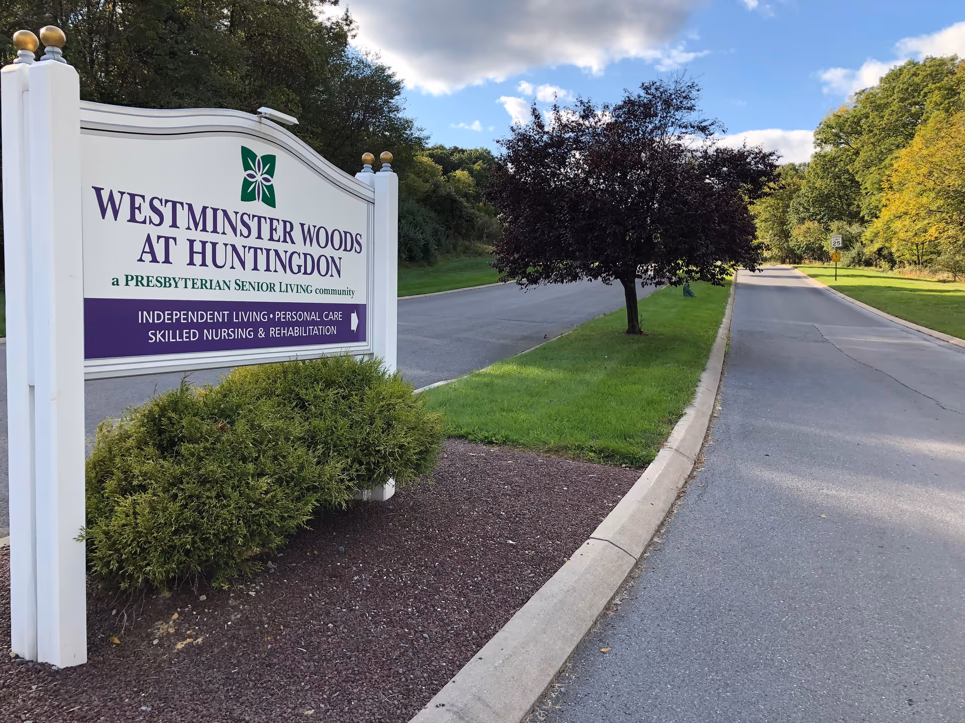 Outdoor view of a road with a sign for Westminster Woods at Huntingdon, a Presbyterian senior living community offering independent living, personal care, skilled nursing, and rehabilitation. The sign is white with purple and green text, surrounded by greenery and trees under a partly cloudy sky.