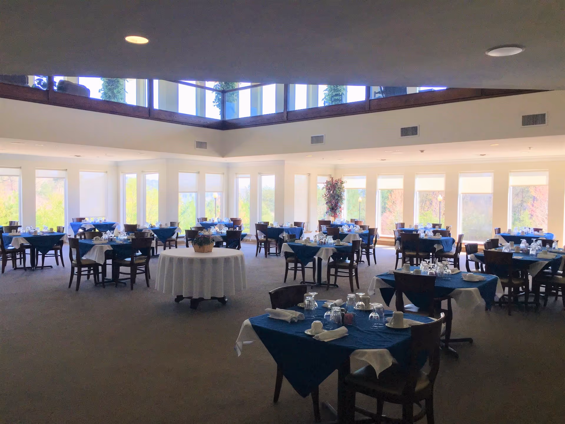 A spacious dining room with multiple tables covered in blue and white tablecloths, each set with cups, glasses, and napkins. Large windows surround the room, allowing natural light to fill the space and providing views of greenery outside. The ceiling has a recessed area with additional windows near the top.