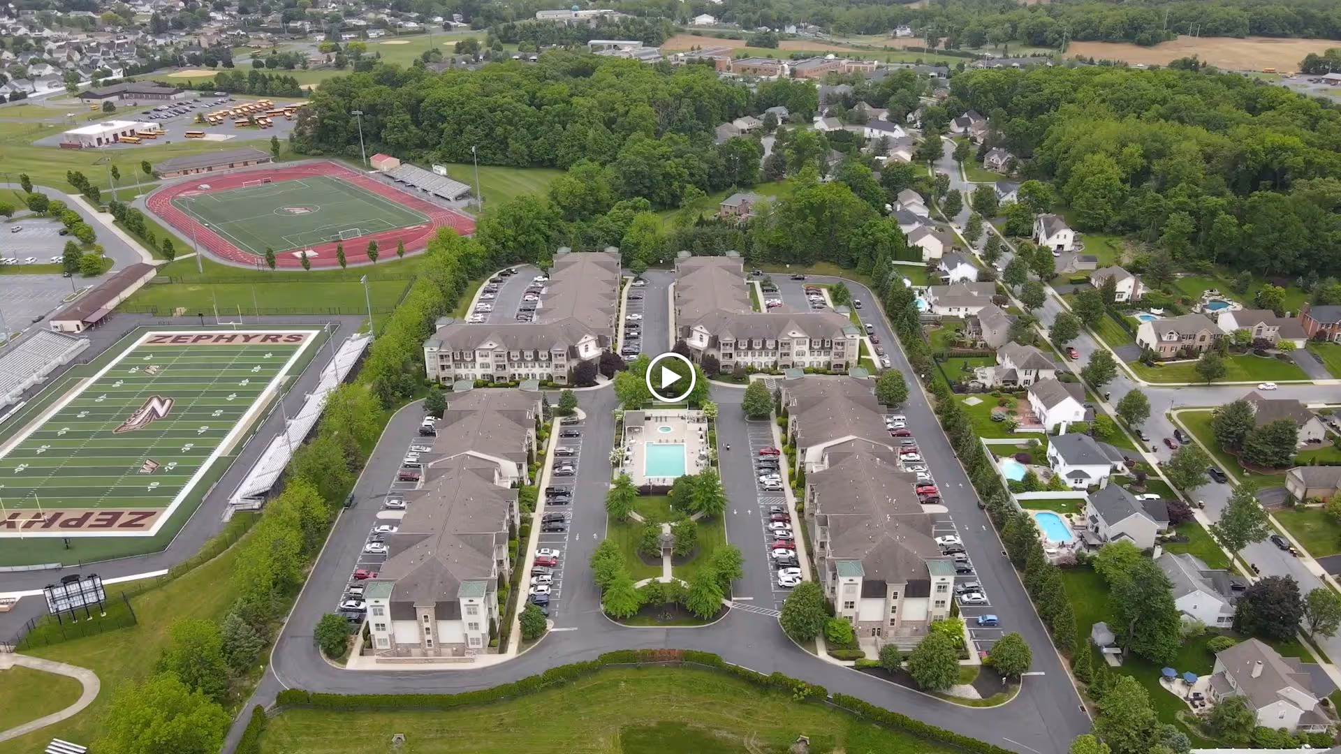 Aerial view of the Horizons at the Village at Whitehall complex showing multiple residential buildings arranged around a central pool, parking lots, and nearby sports fields and houses.