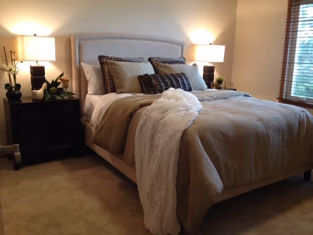 A cozy bedroom with a neatly made bed featuring beige and brown bedding, multiple pillows, and a white throw blanket. Two bedside tables each hold a lamp and decorative items. A window with wooden blinds allows natural light into the room.