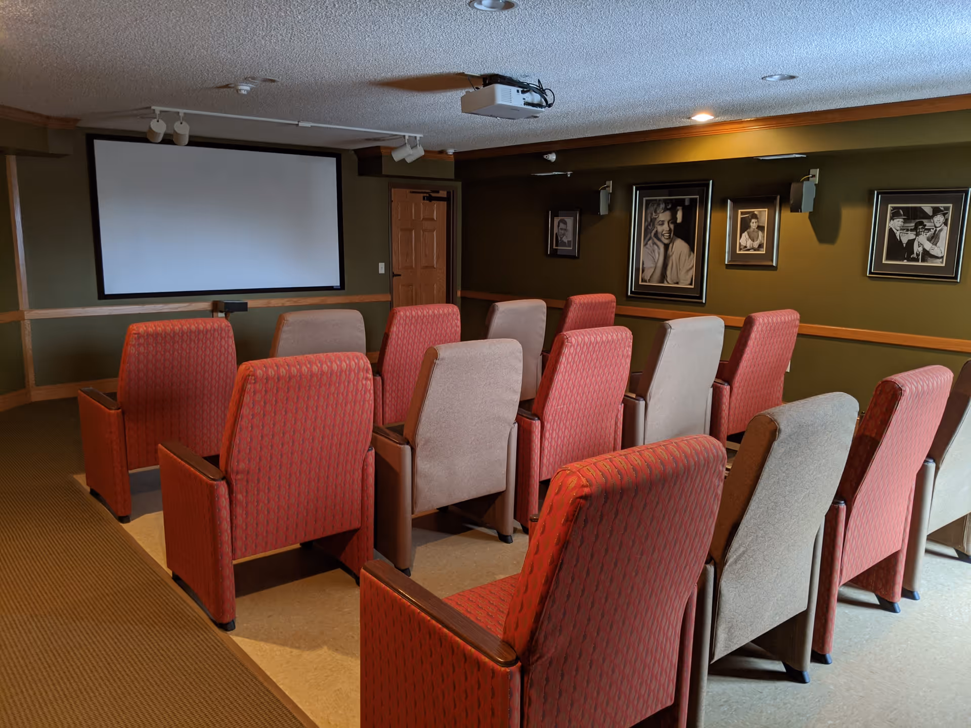 Small theater room with rows of red and beige upholstered chairs facing a large blank projector screen. The walls are olive green with framed black and white photographs, and there is a ceiling-mounted projector above the chairs.