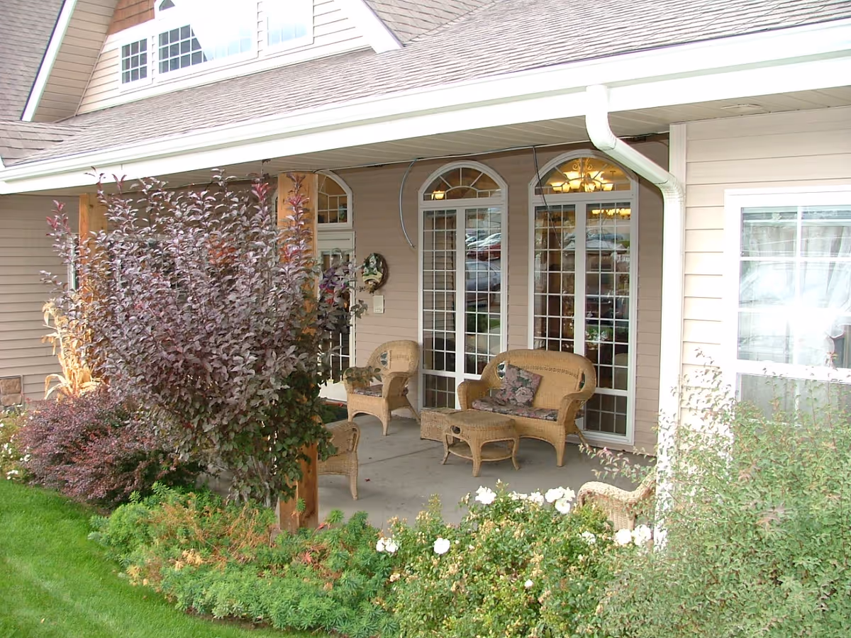 Covered outdoor patio area with wicker furniture including chairs, a loveseat, and a small table. The patio is adjacent to a building with large glass doors and windows. There are various shrubs and flowering plants surrounding the patio.