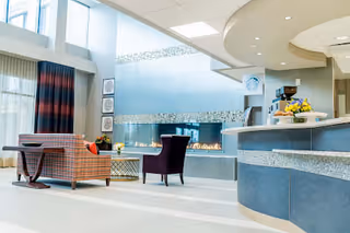 A bright and modern senior living community common area featuring a curved reception desk with a flower arrangement, a long gas fireplace with decorative tiles above it, and comfortable seating including a plaid armchair and a purple chair. Large windows with curtains allow natural light to fill the space.