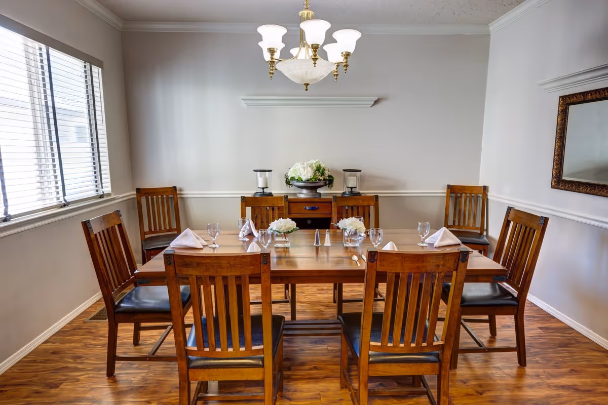A dining room with a wooden table set for six people. The table has six wooden chairs with black cushions, white folded napkins, glassware, and small floral centerpieces. A sideboard with decorative items and a large mirror on the wall are visible. The room has wooden flooring, white walls, and a window with blinds on the left side.