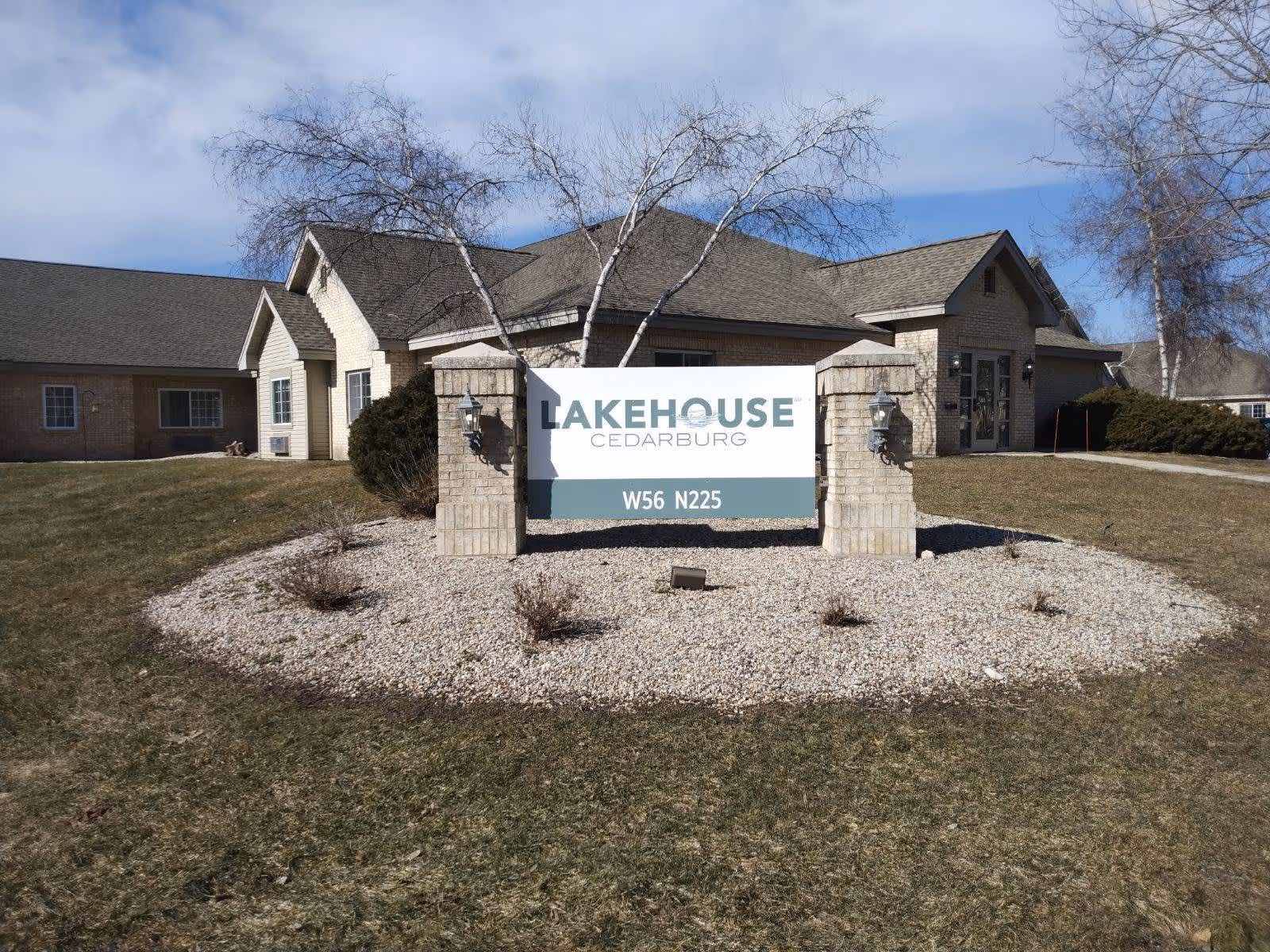 Exterior view of a building with a sign in front that reads 'LakeHouse Cedarburg W56 N225'. The building has beige brick walls, a dark shingled roof, and leafless trees around it. The sign is mounted on two brick pillars surrounded by a bed of white gravel with some small plants.