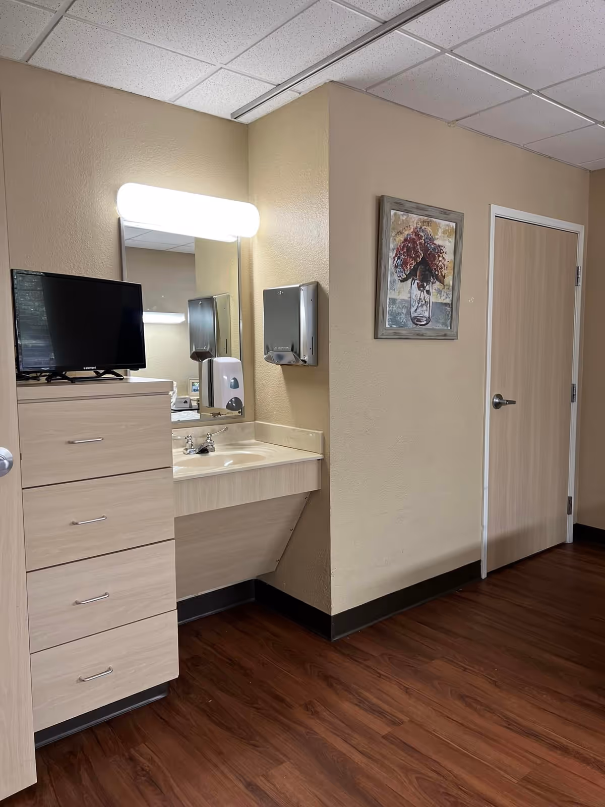 Interior room corner with a sink and mirror under a lit vanity light, a set of drawers with a small TV, wall-mounted dispensers, artwork, and a closed door.