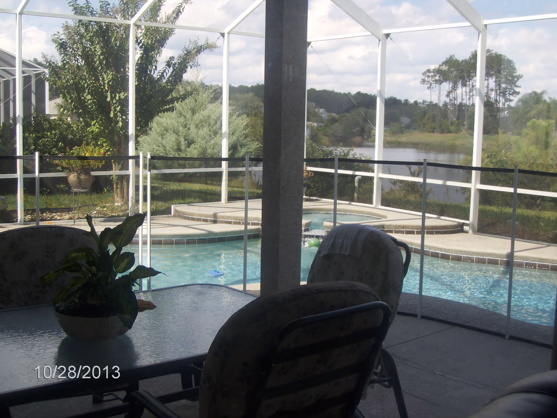Screened patio with a table, chairs and potted plant overlooking a fenced swimming pool and a lake beyond.