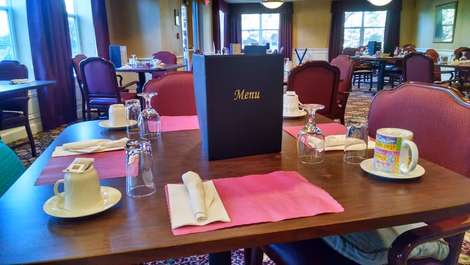 A dining table set for four in a senior living facility dining room. The table has pink placemats, folded napkins, upside-down glasses, coffee cups on saucers, and a black menu standing upright in the center. The room has multiple tables and chairs with red upholstery, large windows with curtains, and a patterned carpet.