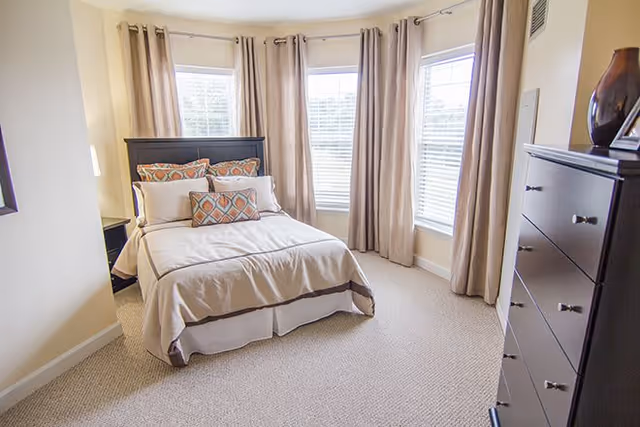 A bright bedroom with a neatly made bed featuring beige and brown bedding and decorative pillows. The room has three large windows with beige curtains, beige walls, and a carpeted floor. There is a dark wooden dresser with a vase and picture frame on top to the right of the bed.