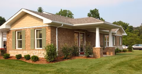 Exterior view of a single-story brick and stone building with a covered porch, surrounded by green grass and shrubs under a clear sky.