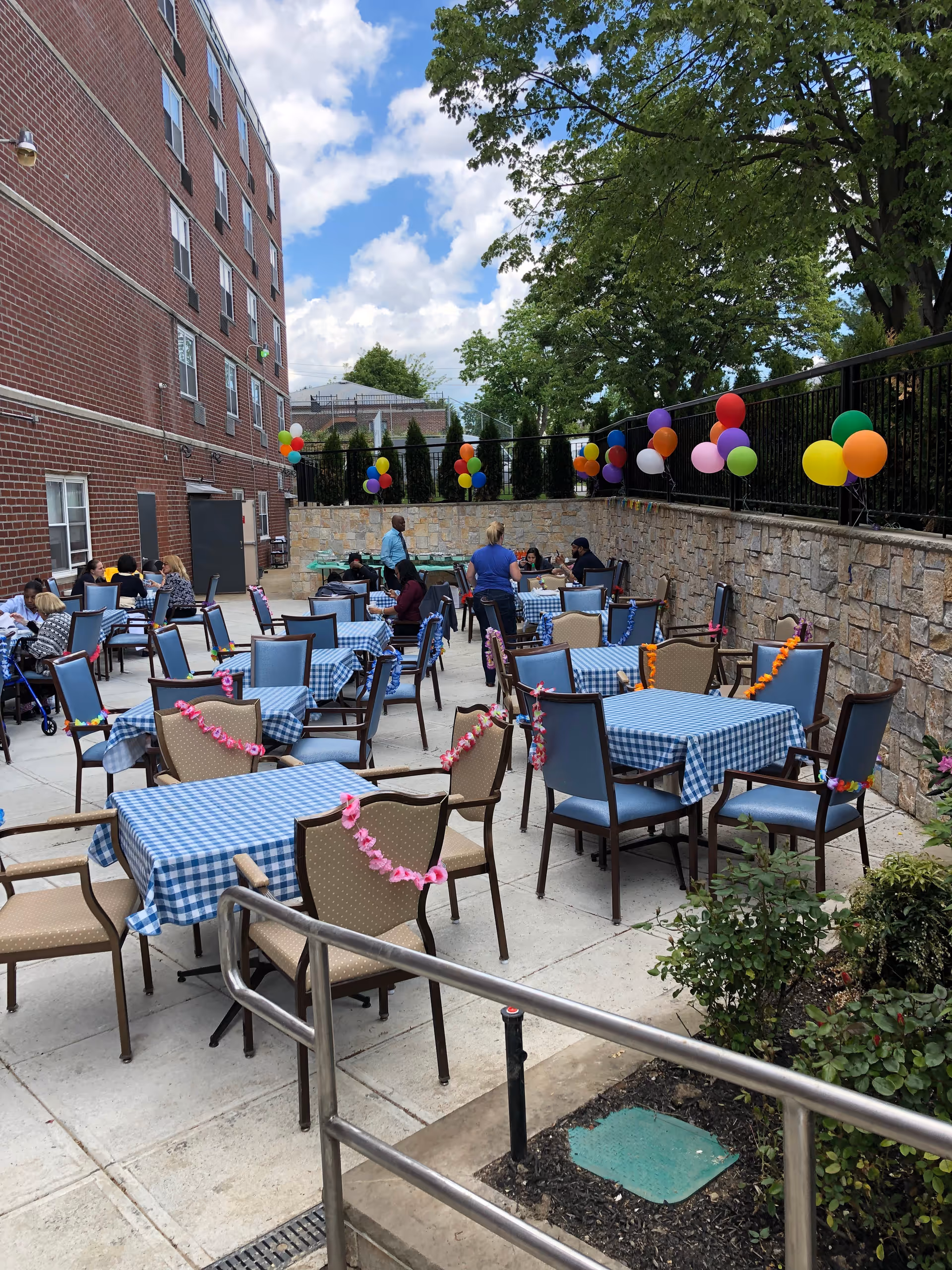 Outdoor patio at a nursing facility with multiple tables covered in blue checkered tablecloths, chairs decorated with leis, balloons along a stone wall, and people gathered.