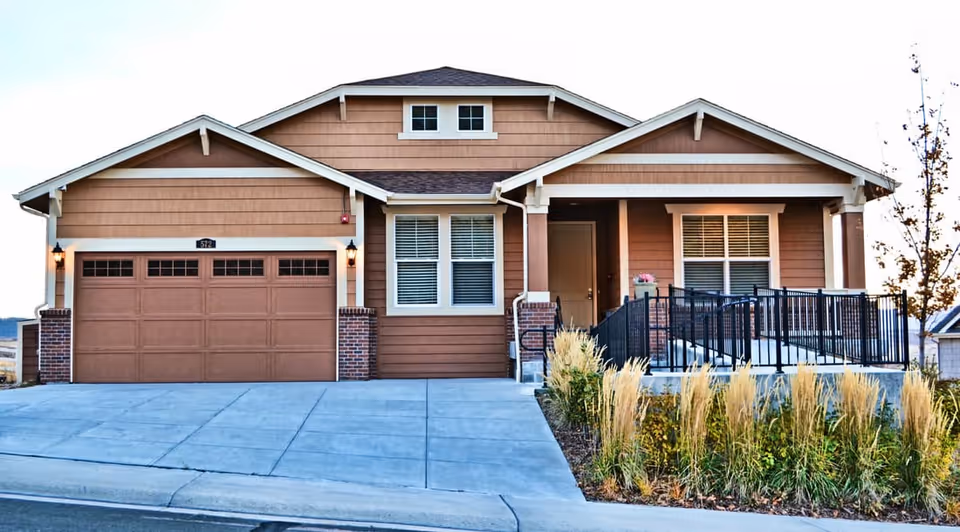 Exterior front view of a single-story residential building with a two-car garage, a small porch with a ramp and railing, and landscaping with tall grasses and plants along the driveway.