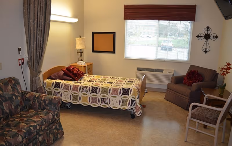 A cozy room with a single bed covered with a patterned quilt, a bedside table with a lamp, a window with blinds and a red valance, a cushioned armchair with a red pillow, a patterned couch, and a small table with a plant. The room has beige walls and flooring.