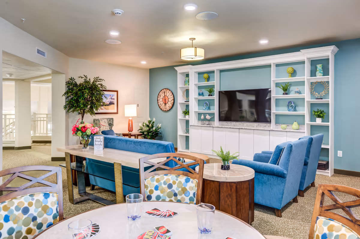 A bright and cozy living room area in a senior living facility featuring blue upholstered chairs and a sofa, a large flat-screen TV mounted on a white built-in shelving unit with decorative items, a round table with patterned chairs and UNO cards, and various plants and wall decorations including a clock and framed picture.