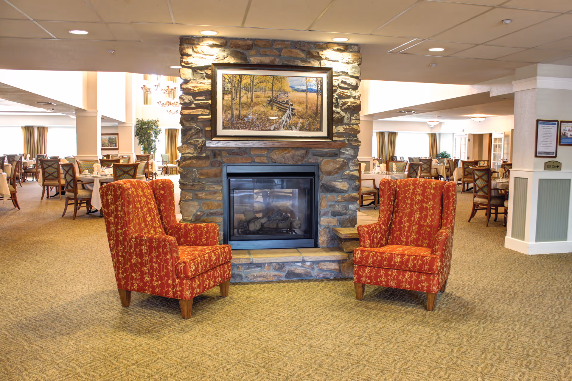 Interior view of a senior living facility dining area with a stone fireplace in the center. Two red patterned armchairs are placed facing each other in front of the fireplace. Behind the fireplace, there are multiple dining tables with chairs, set with tablecloths and napkins. The room is well-lit with ceiling lights and natural light from windows.