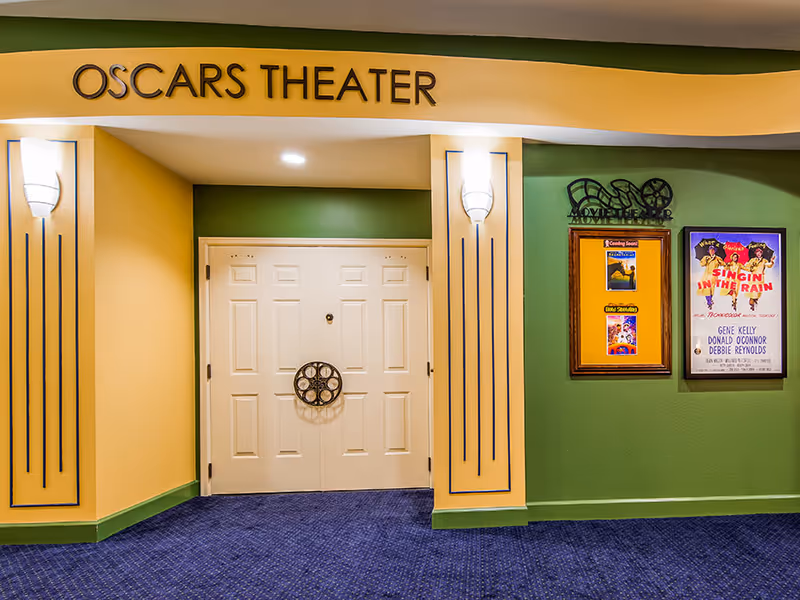 Entrance to Oscars Theater with double white doors, yellow and green walls, blue carpet, wall sconces, and movie posters including Singin' in the Rain.