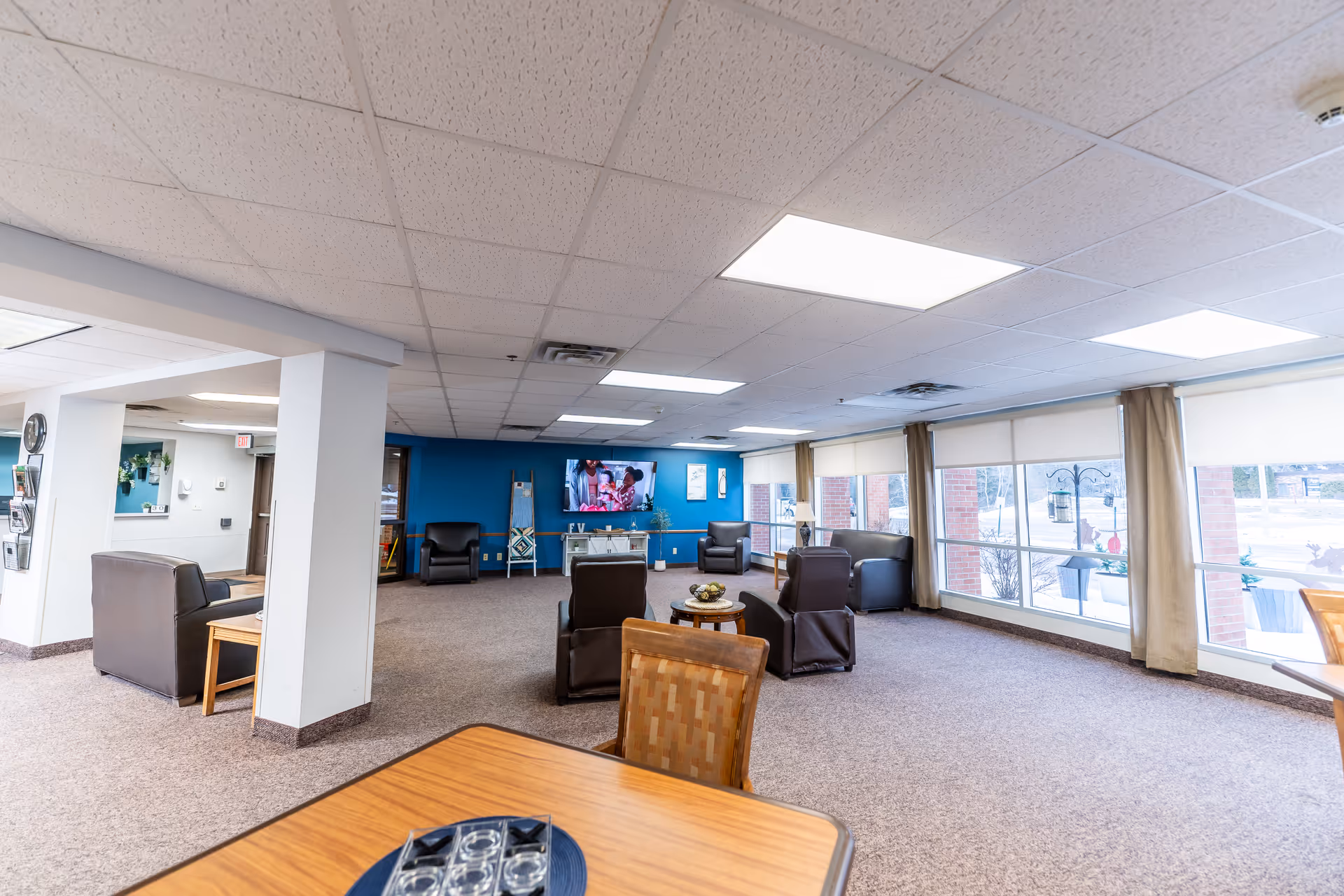 Spacious communal lounge with chairs, tables, a wall-mounted TV, and large windows letting in daylight.