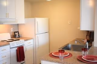 A small kitchen area with white cabinets, a white refrigerator, an electric stove with a red towel hanging on the handle, and a countertop with a sink. Two place settings with red plates and glasses are arranged on the countertop.
