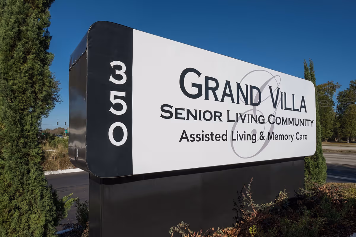 Outdoor sign for Grand Villa Senior Living Community, offering Assisted Living and Memory Care, with the address number 350 displayed vertically on the left side of the sign. The sign is surrounded by greenery and set against a clear blue sky.