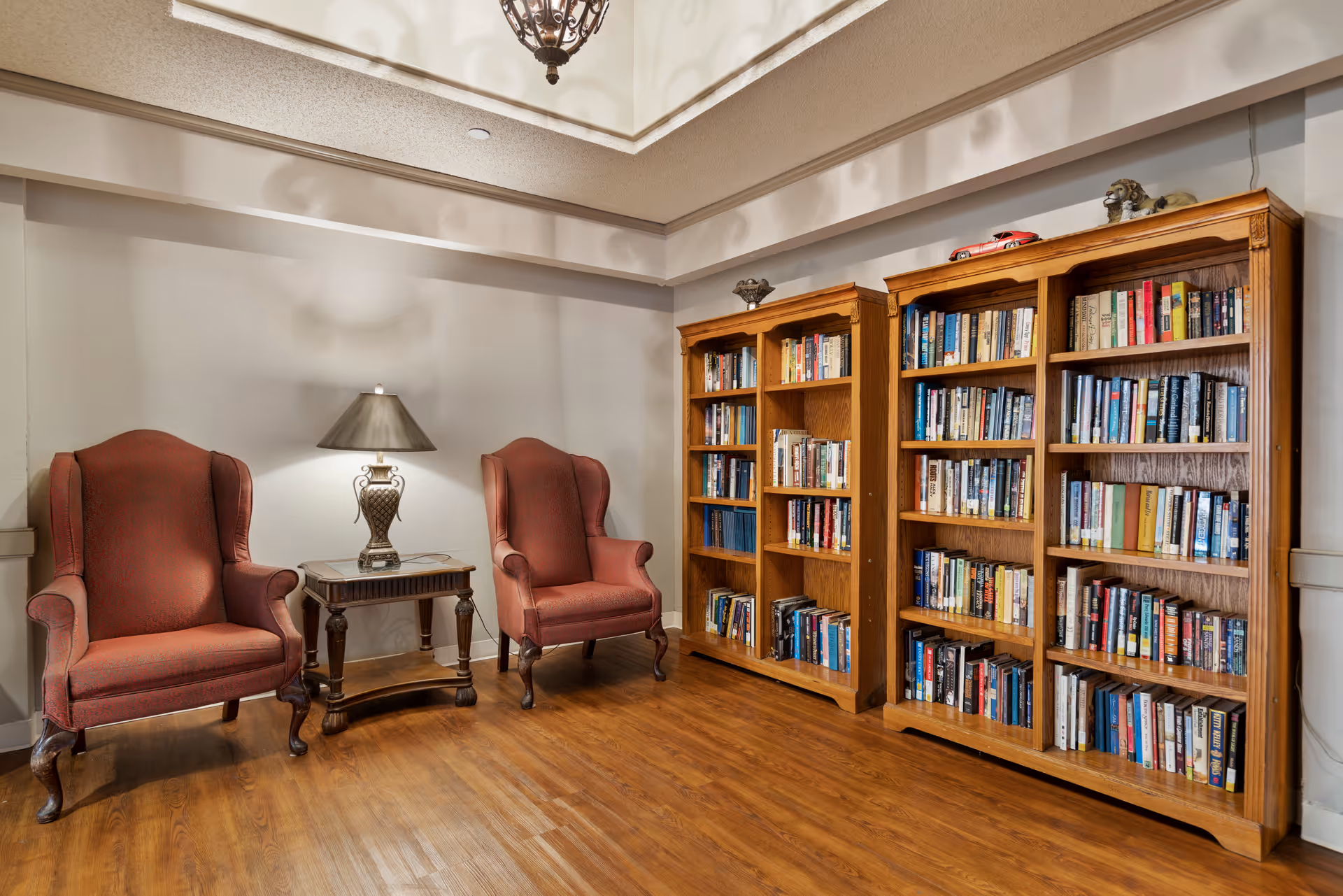 A cozy reading area with two red upholstered armchairs separated by a wooden side table with a decorative lamp. To the right, there are three wooden bookshelves filled with books. The room has wooden flooring and a high ceiling with a hanging light fixture.