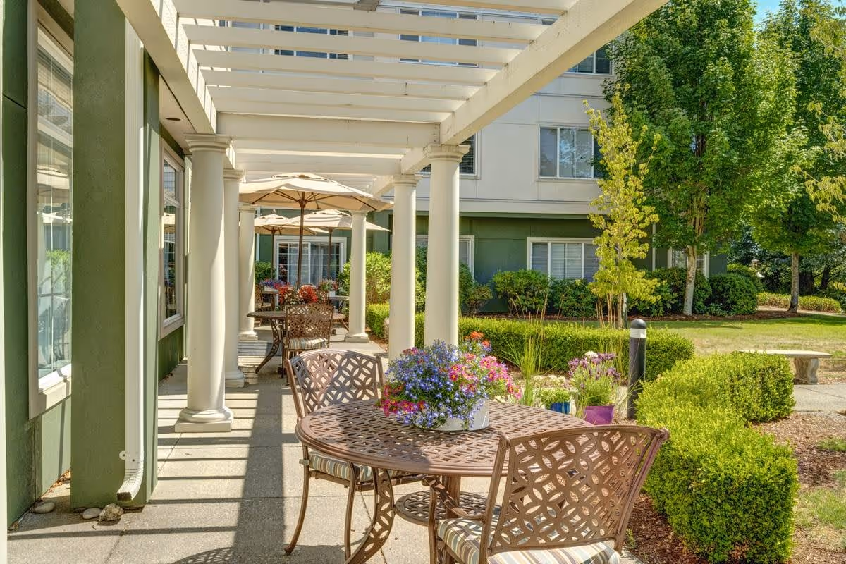 Outdoor patio area at Cordata Court with metal tables and chairs featuring intricate designs, potted flowers on the tables, white pergola overhead, and green bushes and trees surrounding the space.