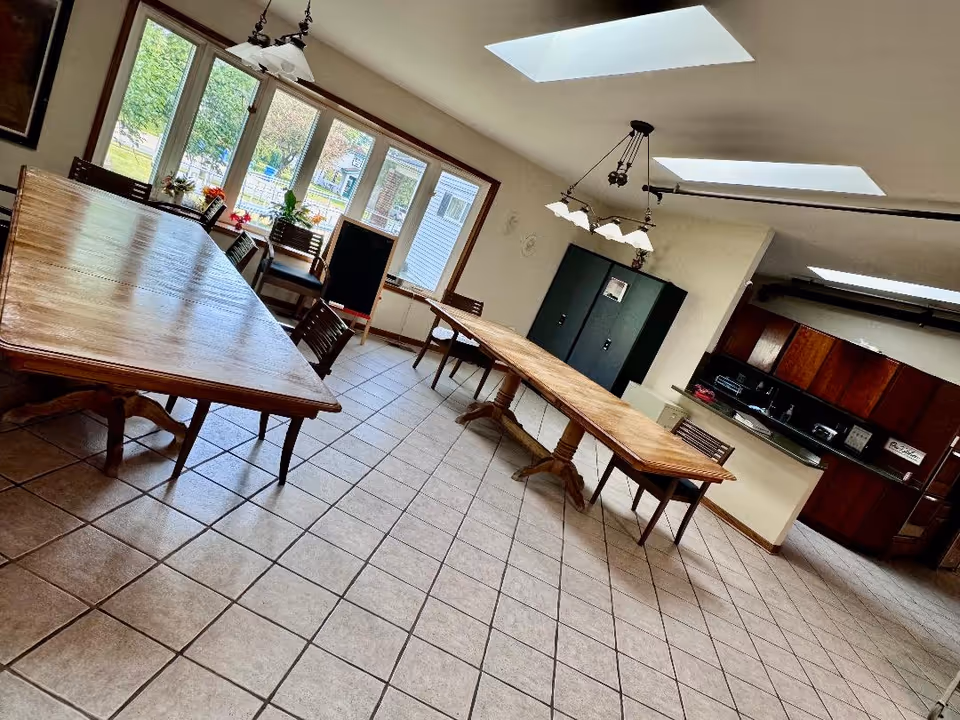 Bright communal dining room with long wooden tables and chairs, skylights, and a small kitchen area in the back.