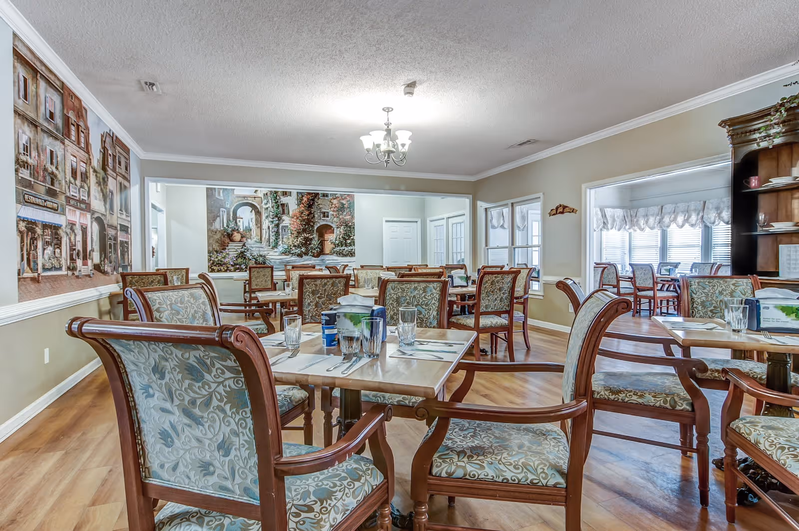 A dining room in a senior living facility with multiple wooden tables and upholstered chairs arranged neatly. The tables are set with glasses, napkins, and utensils. The walls feature large murals depicting scenic street views, and there is a chandelier hanging from the ceiling. The room has wood flooring and large windows allowing natural light.