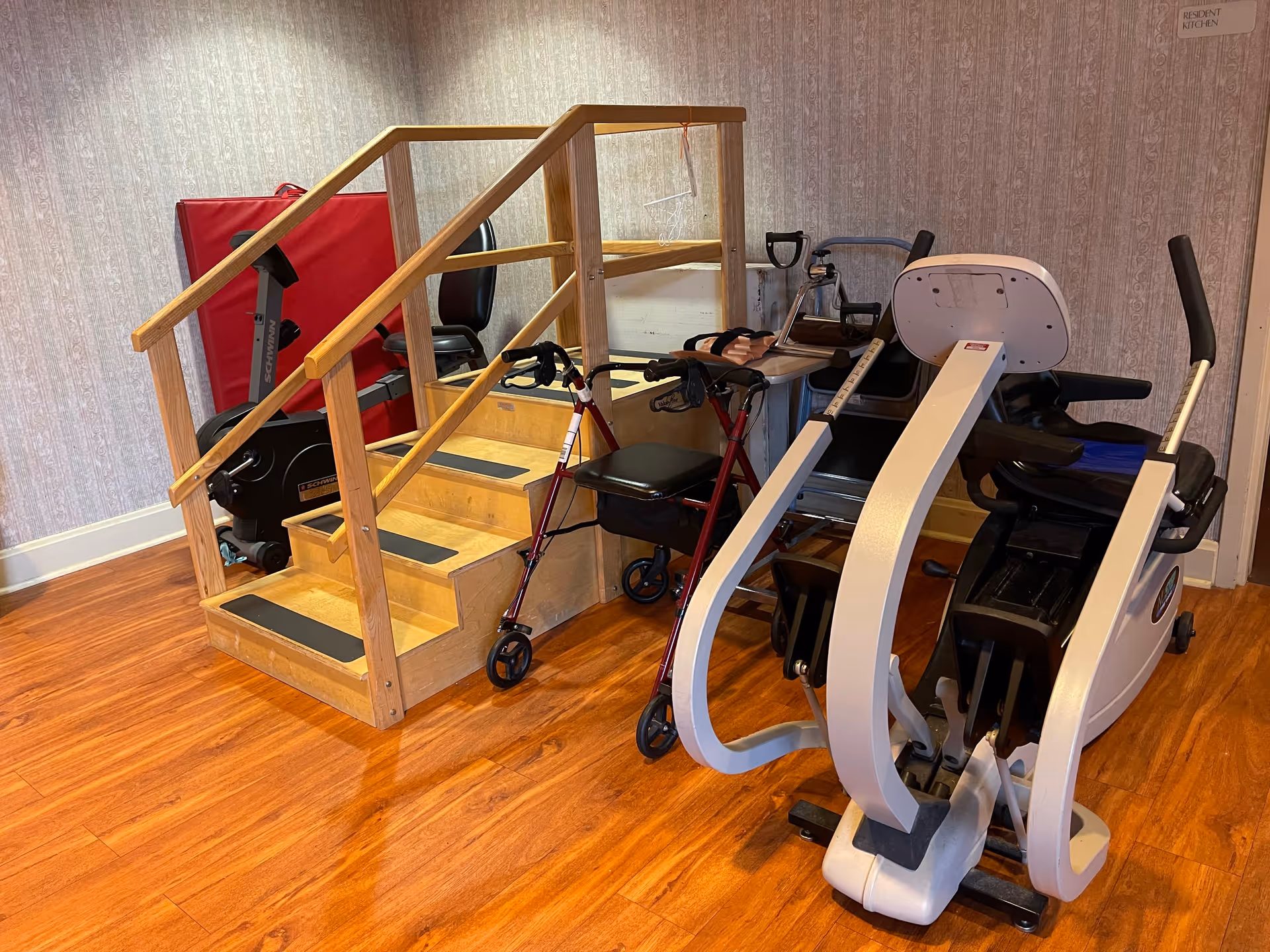 Indoor exercise area with wooden stairs equipped with handrails, a red walker, a stationary exercise bike, and other exercise equipment on a wooden floor with wallpapered walls.