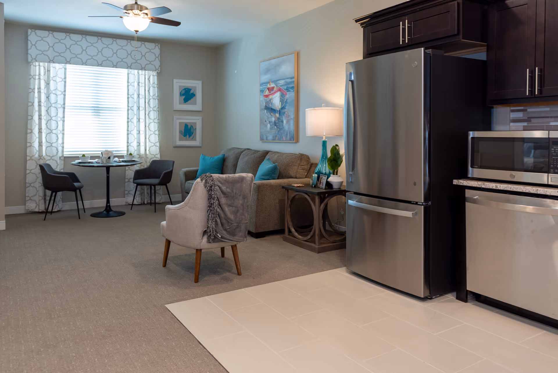 Interior view of a senior living facility showing a combined living room and kitchen area. The living room has a gray sofa with teal pillows, a beige armchair with a gray throw, a side table with a lamp and decorative items, and a painting of a boat on the wall. The kitchen area features stainless steel appliances including a refrigerator, microwave, and dishwasher, with dark wood cabinets. In the background, there is a small dining area with a round table and two chairs near a window with patterned curtains.