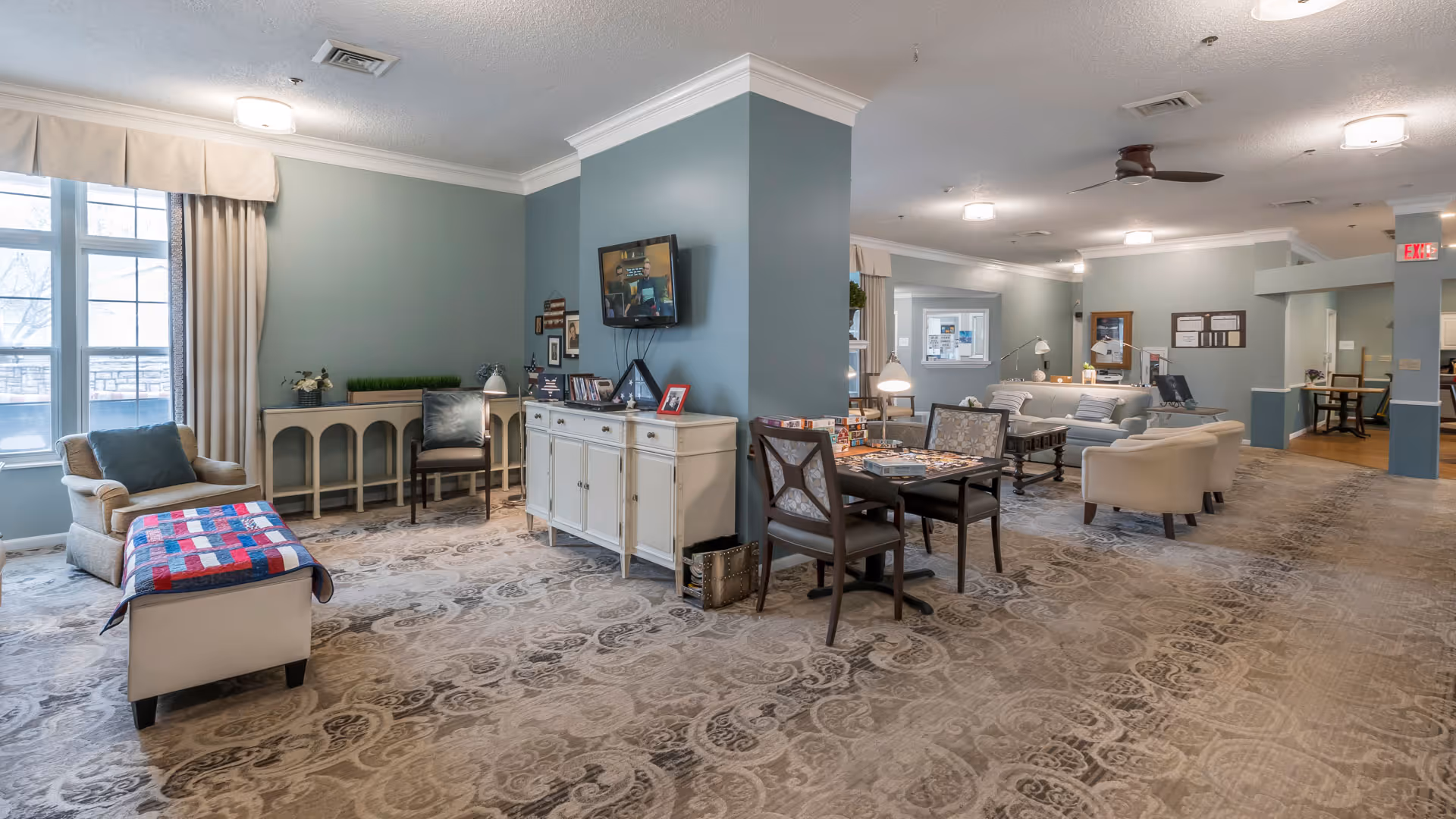 A spacious and well-lit common area in a senior living facility with comfortable seating including armchairs and sofas, a table with chairs set up for board games, a wall-mounted TV, and large windows with curtains allowing natural light to enter. The room has light blue walls, patterned carpet, and ceiling lights with a ceiling fan.
