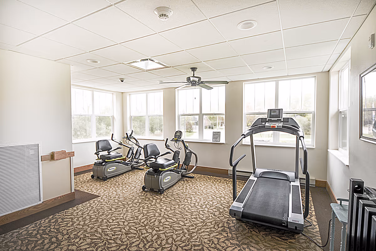 A bright exercise room with large windows letting in natural light. The room contains two recumbent stationary bikes and one treadmill on a patterned carpet floor. A ceiling fan is mounted in the center of the white tiled ceiling.