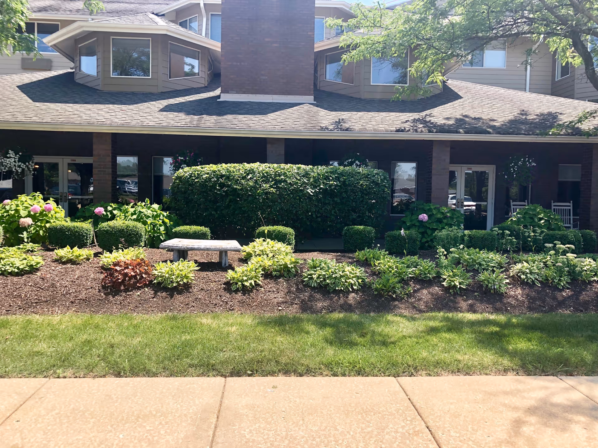 Front exterior view of Solstice Senior Living at Joliet showing a landscaped garden with various green shrubs, flowering plants, and a stone bench in front of the building. The building has a brick facade with large windows and a covered porch area with rocking chairs.