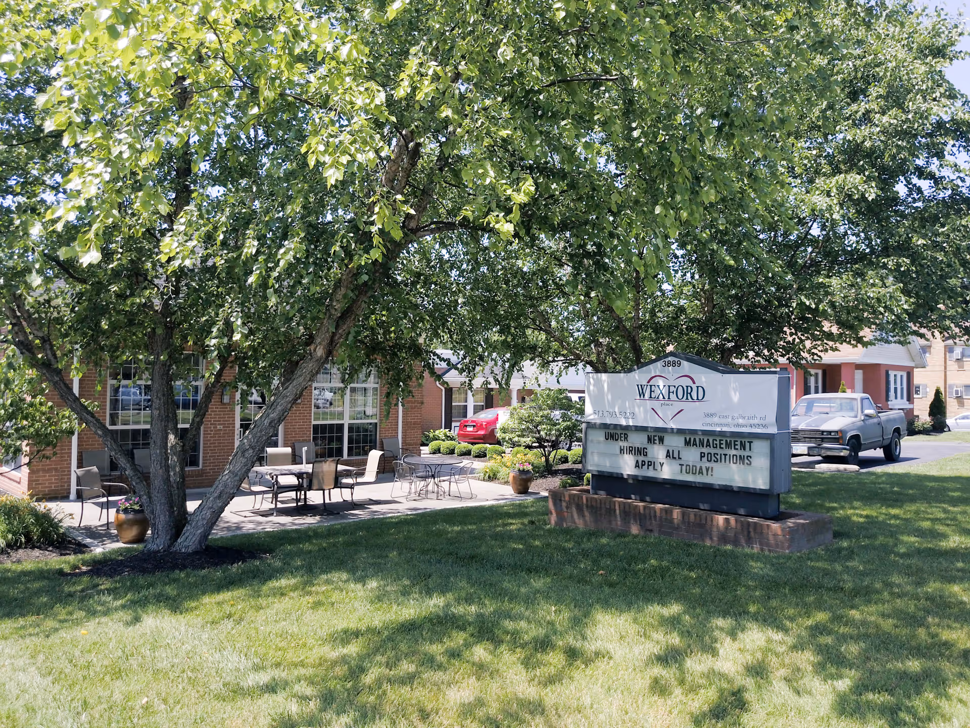 Outdoor view of a senior living facility with green grass, large leafy trees, and a brick building with large windows. There are several patio tables and chairs on a paved area near the building. A sign in front of the building reads 'Wexford Place' and advertises that the facility is under new management and hiring all positions.