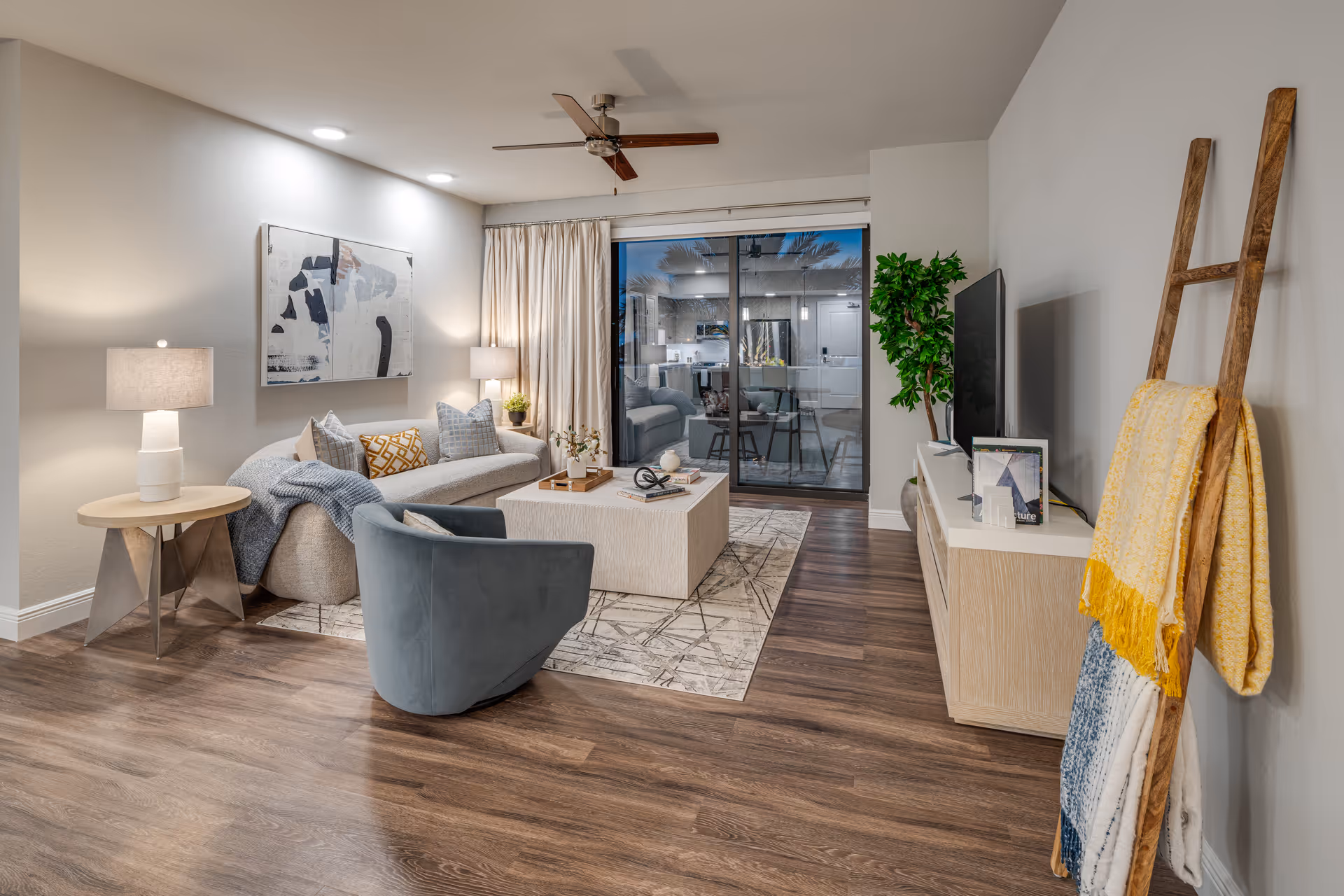 A modern living room with wooden flooring, a gray sofa with multiple cushions, a blue armchair, a square coffee table on a patterned rug, two side tables with lamps, a ceiling fan, a large flat-screen TV on a wooden console, a decorative ladder with blankets, and a large sliding glass door leading to another room.