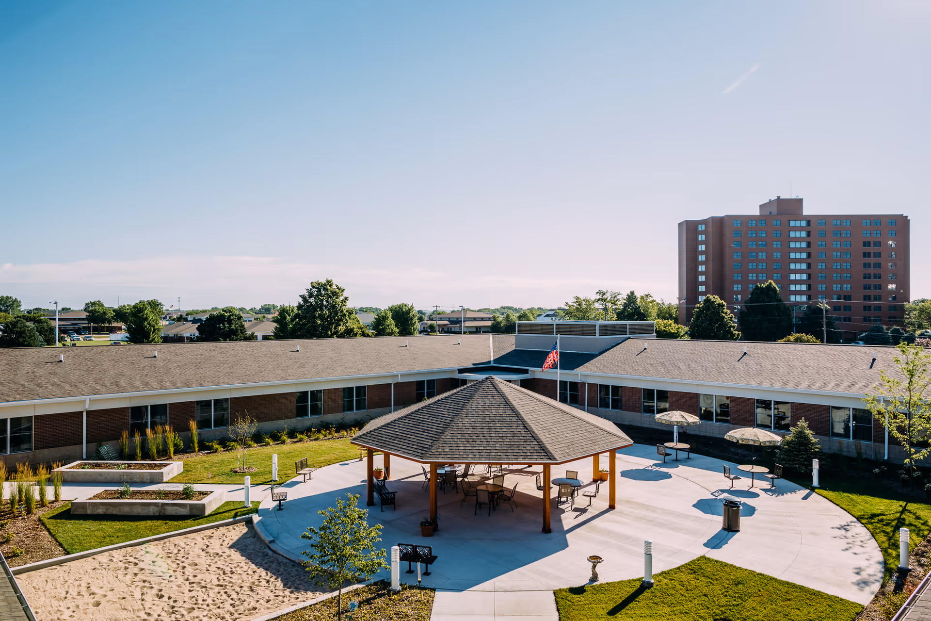 Outdoor courtyard area of a senior living facility with a central gazebo surrounded by tables and chairs, landscaped garden beds, and a large building in the background under a clear blue sky.