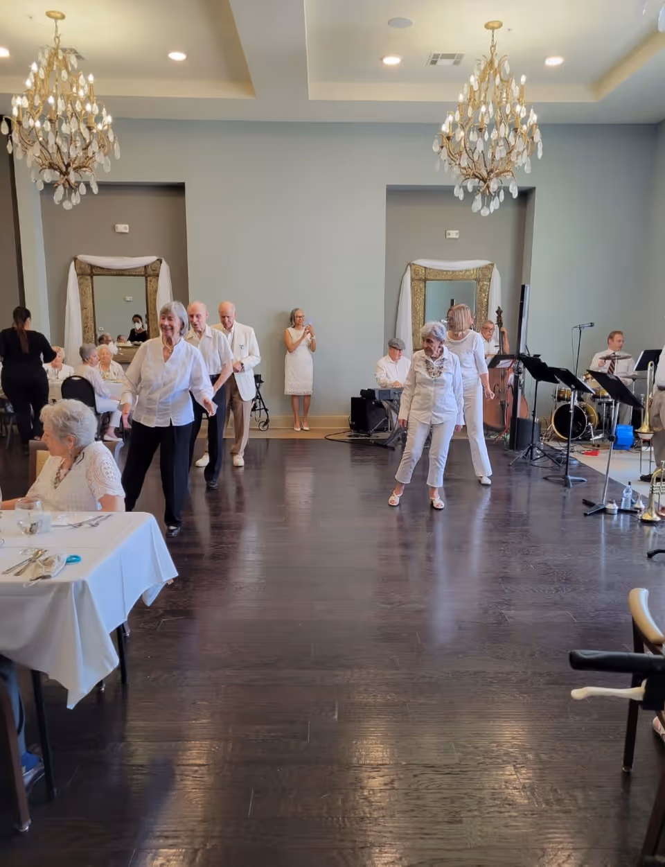 A group of elderly people dancing and socializing in a spacious room with dark wooden floors and light-colored walls. There are chandeliers hanging from the ceiling, and a live band playing music in the background. Some people are seated at tables with white tablecloths, while others are standing or dancing. A woman in a white dress is taking a photo in the background.