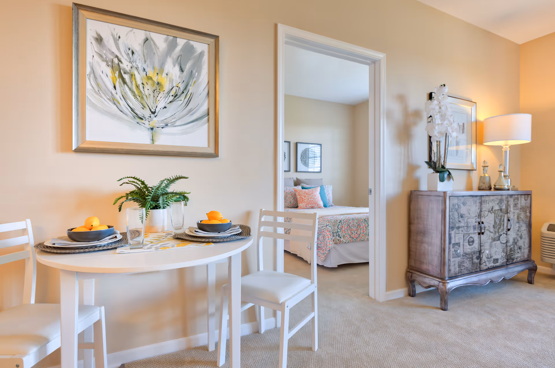 A cozy dining area with a small white round table set for two with bowls of oranges and glasses. A green plant is placed in the center of the table. The room has beige walls and carpeted floor. A framed floral painting hangs above the table. Through an open doorway, a bedroom with a bed covered in a colorful quilt and pillows is visible. To the right, there is a decorative wooden cabinet with a lamp, framed picture, and a white orchid on top.