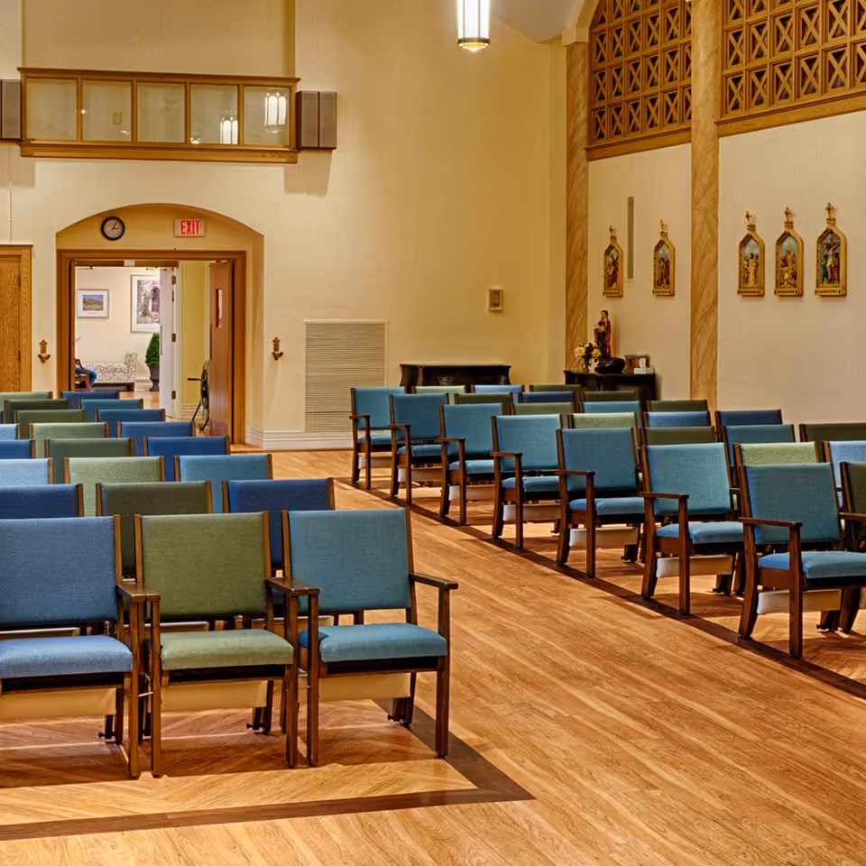 Interior view of a chapel or meeting room with rows of wooden chairs upholstered in blue and green fabric. The room has light-colored walls, wooden flooring, religious artwork on the walls, and a statue on a table at the far end. There is an exit door with a red exit sign above it and a clock on the wall.