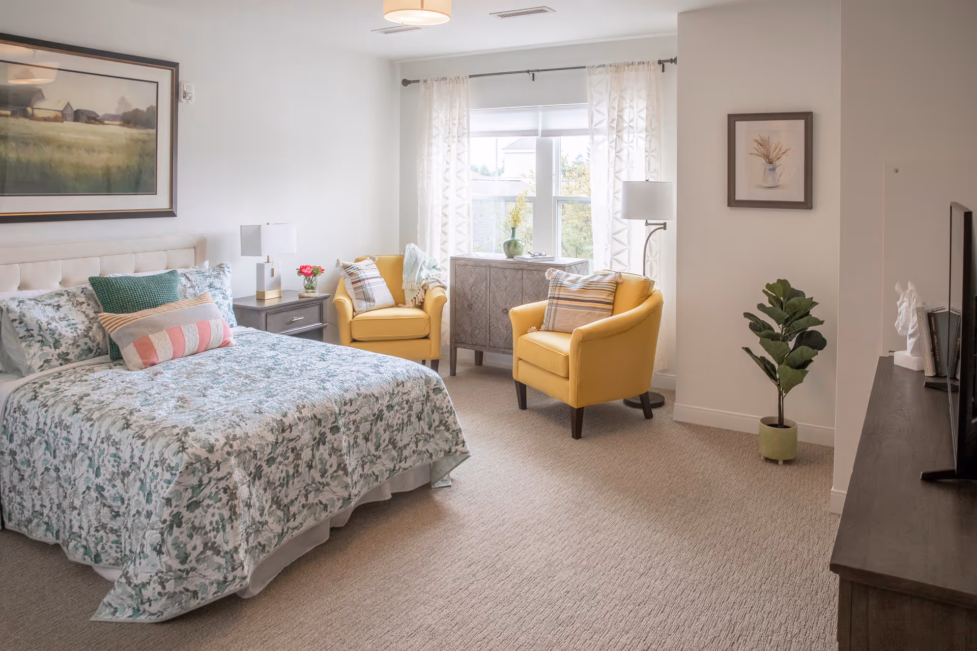 A bright furnished bedroom with a bed featuring floral bedding, two yellow armchairs by a window, bedside tables and a TV on a dresser.